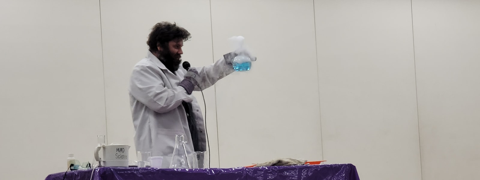 Man in la white lab coat speaking into a mic and holding a glass container with a blue liquid.