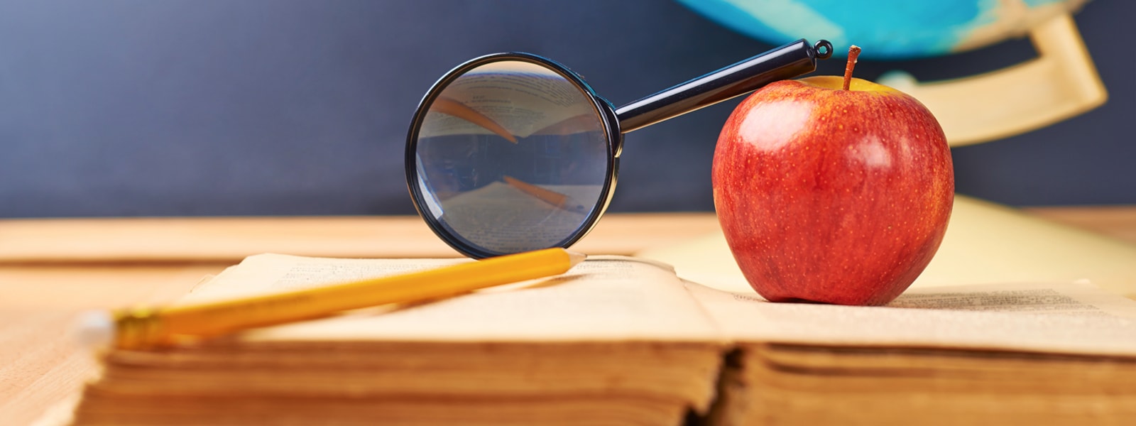 An apple, a pencil and magnifying glass on top of a book.