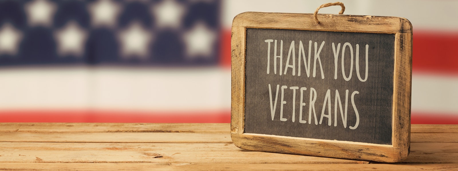 A chalk board sign saying Thank You Veterans sitting on a wood table with flag in the background.