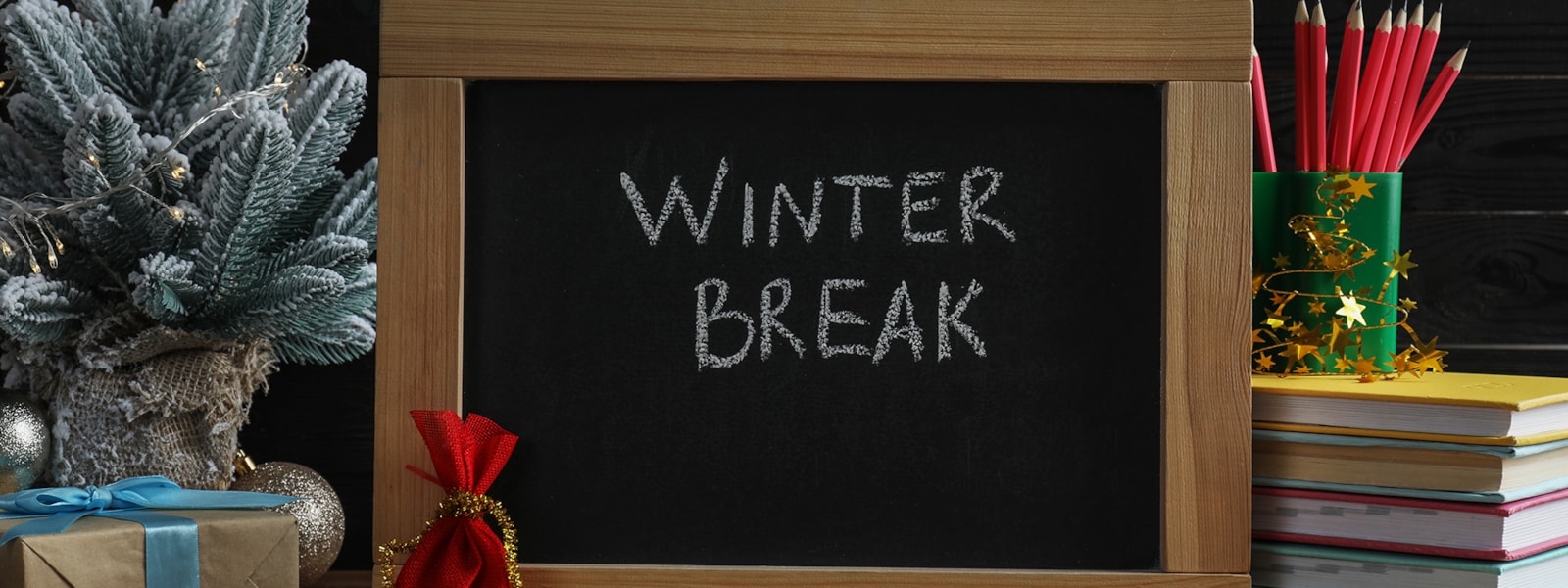 A blackboard showing Winter Break. Surrounded by presents, books, pencils, and a small tree
