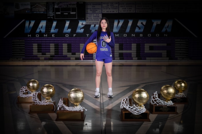 Girls basketball player standing on court with ball surrounded by state trophies.