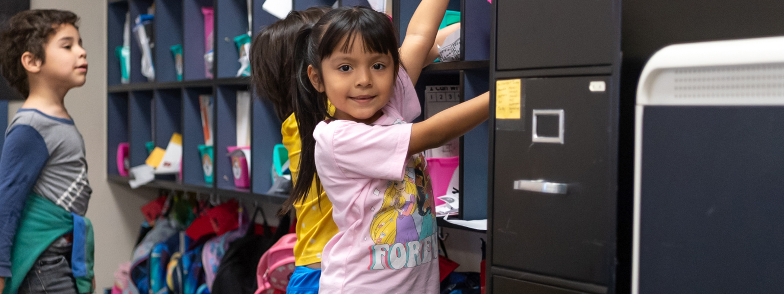Kindergarten student reaching into a cubby whille smiling at the camera