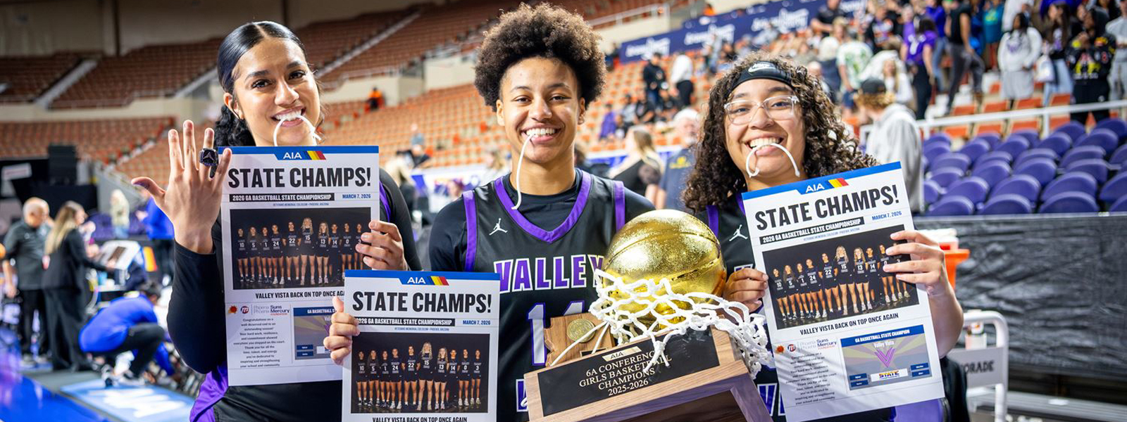 Three girls basketball players hold up novelty newspapers with the headline, "State Cahmps!" while the player in the middle also holds the 6A Conference Girls Basketball Champion trophy 
